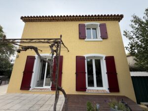 Maison jaune avec volets rouges et pergola.
